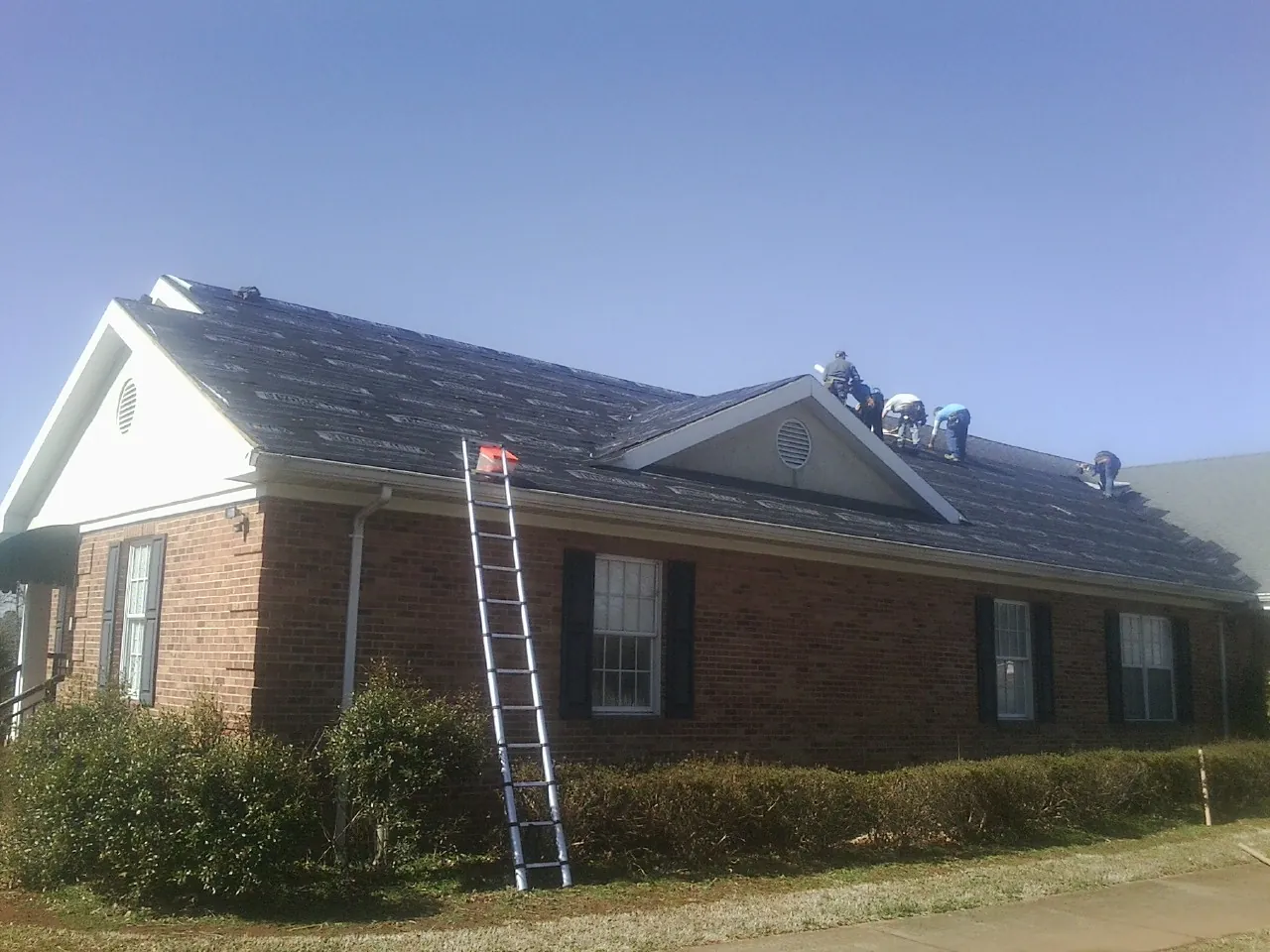 Crew tearing shingles off a brick ranch roof with a ladder set — Emergency Roof Repair work by our crew in Coram