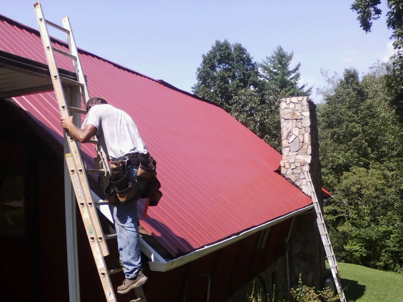 Roofer on a ladder beside a red metal roof and stone chimney — Soffit & Fascia Repair by Roofing Brothers in Coram